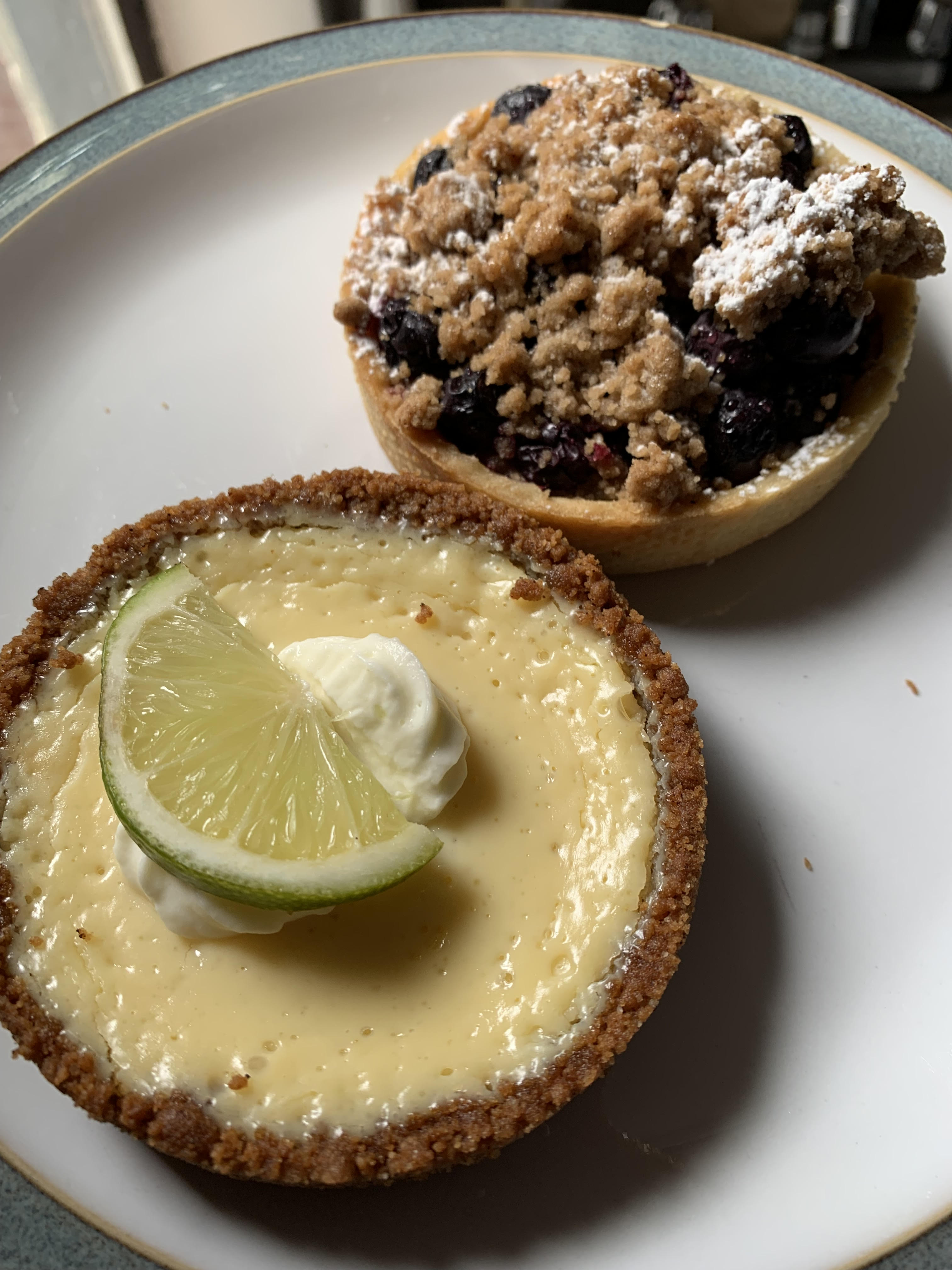 Two tarts, one key lime topped with a lime slice and whipped cream, one blueberry topped with oatmeal crumble and powdered sugar.