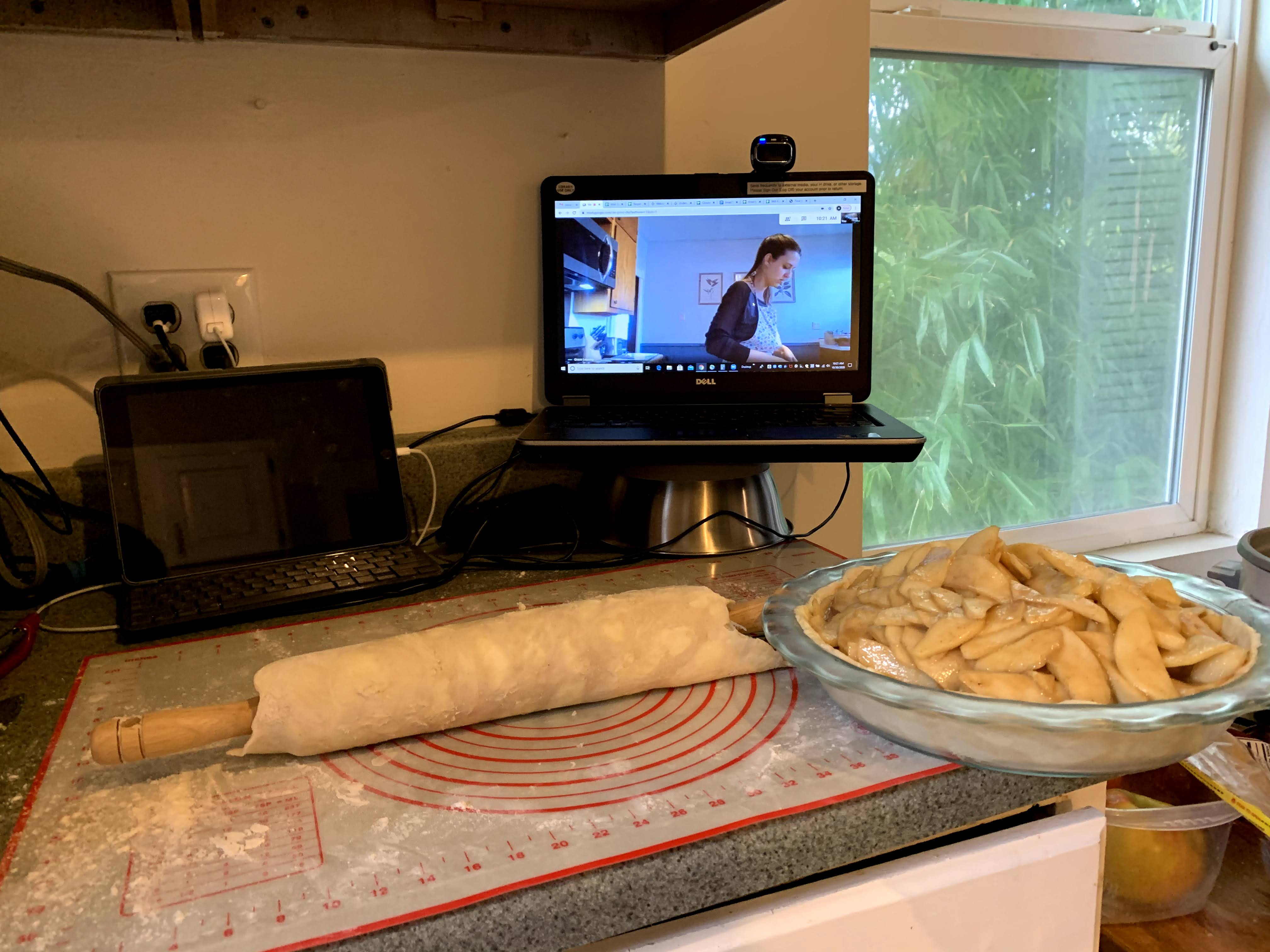 An apple pie in progress, sprawled across a counter. There is a computer propped up with another baker seen over Zoom.